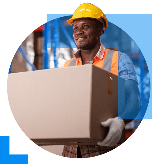 A man in work clothing and a yellow hard hat, carrying a large parcel in a warehouse setting, furfilling an online order, representing the E-commerce sector.