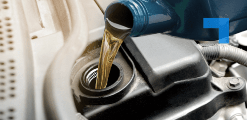 Motor oil is being poured from a blue plastic container into the oil fill port of a car engine. The image captures a close-up view of the engine bay during an oil change, emphasizing the golden-brown liquid flowing smoothly from the container into the vehicle.