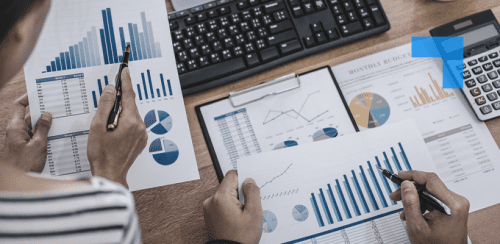 Two individuals reviewing financial documents at a desk equipped with a keyboard, calculator, and clipboard. The documents feature colourful bar graphs, pie charts, and a line graph illustrating yearly revenue, expenses, profit, and monthly budget trends, indicating a focused financial analysis session.