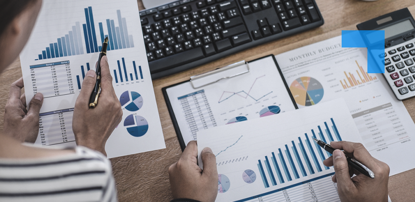 Two individuals reviewing financial documents at a desk equipped with a keyboard, calculator, and clipboard. The documents feature colourful bar graphs, pie charts, and a line graph illustrating yearly revenue, expenses, profit, and monthly budget trends, indicating a focused financial analysis session.
