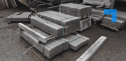 Stacked concrete blocks or beams secured with green straps and placed on wooden pallets in an outdoor storage area. Various sizes are visible, some with markings or labels. Additional stacks and metal structures appear in the background, suggesting a construction site or materials yard.