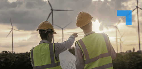 Two workers in yellow hard hats and high-visibility vests stand in front of wind turbines under a cloudy sky with the sun partially visible. One holds a blueprint while the other points toward the turbines, suggesting a discussion or inspection.