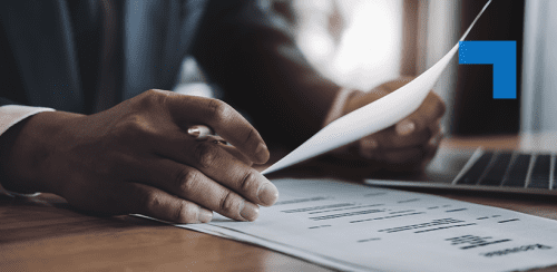 A person in a business suit sitting at a desk, reviewing printed documents with one hand holding a paper and the other resting on a stack of reports. A laptop is partially visible on the desk, with natural light coming from a window in the background.