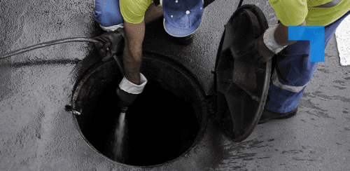 Two workers wearing high-visibility clothing open a manhole cover and use a hose to clean or inspect the inside of the sewer.