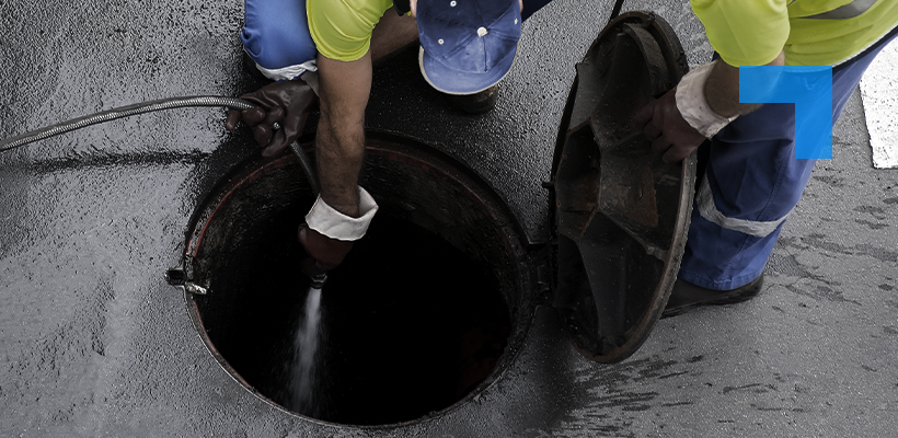 Two workers wearing high-visibility clothing open a manhole cover and use a hose to clean or inspect the inside of the sewer.