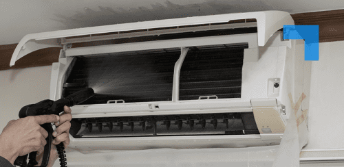A wall-mounted split air conditioner with its front cover open while a person uses a spray nozzle to clean the internal coils.