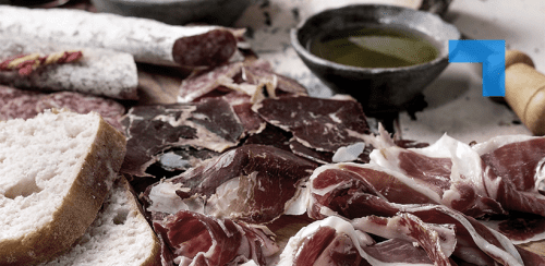 Assorted cured meats sliced and arranged on a wooden surface alongside crusty bread, salami, and a small bowl of olive oil, presented as a rustic charcuterie spread.