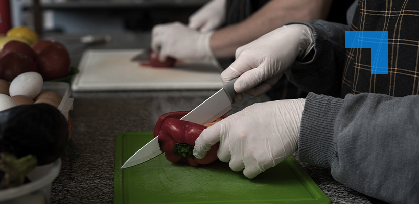 Hands of a person chopping a vegetable with a sharp knife while wearing white gloves for hygiene purposes.