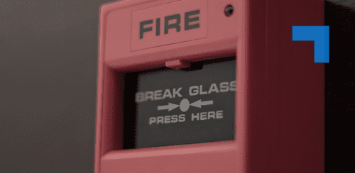 Close-up of a red wall-mounted fire alarm call point displaying the words “FIRE” and “BREAK GLASS – PRESS HERE.”