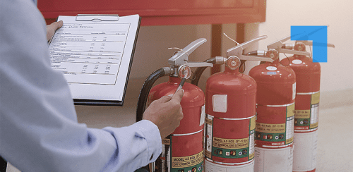 Fire safety technician carrying out inspection of multiple fire extinguishers with checklist on clipboard.