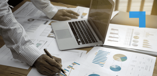A person in a striped shirt reviews printed financial charts and graphs spread across a desk while using a laptop and marking data with a pen, suggesting analysis, reporting, or performance review work.
