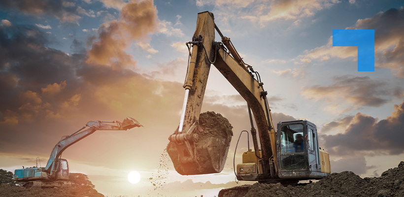 Two excavators working at a construction site at sunset, with the larger machine in the foreground lifting a bucket of soil while a smaller excavator operates in the background under a cloudy sky.