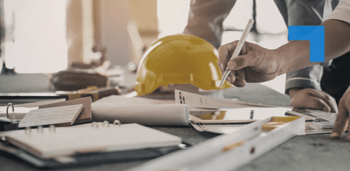 Close-up of a person marking construction plans on a desk with a pen, with a yellow hard hat, ruler, and notebooks visible in a construction workspace.