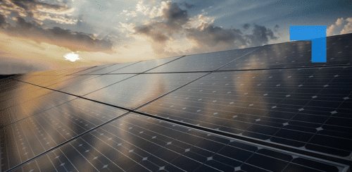 Solar panels arranged in rows under a partly cloudy sky at sunset, reflecting sunlight, with warm orange and blue tones across the scene.