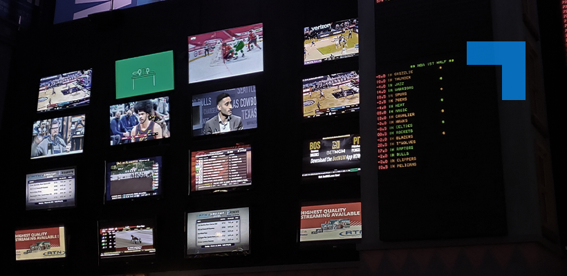 A wall of multiple TV screens in a dimly lit sportsbook or control room, each displaying different live sports events and broadcasts, alongside digital betting odds boards with scrolling numbers and team names.