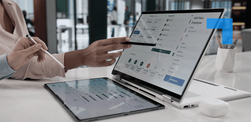 A professional using a tablet and touchscreen laptop displaying HR and workforce management software dashboard in a modern office setting.