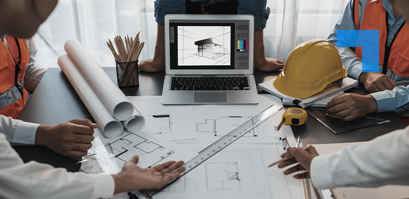 Architects and engineers reviewing building plans around a desk with a laptop displaying a 3D model, alongside blueprints, measuring tools and a hard hat, collaborating on a construction design project.