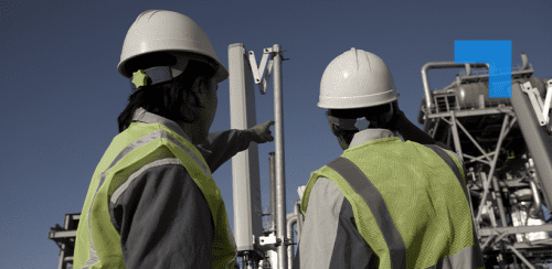 Two engineers in hard hats and high-visibility vests inspect industrial equipment at an outdoor facility, with one pointing toward a vertical structure.