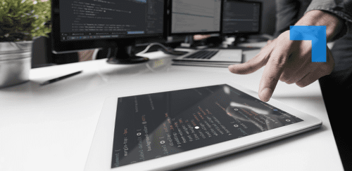 A person points at a tablet displaying lines of programming code on a desk beside multiple computer monitors showing software development screens in an office workspace.