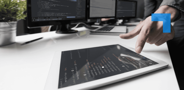A person points at a tablet displaying lines of programming code on a desk beside multiple computer monitors showing software development screens in an office workspace.