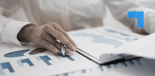A close-up of a person’s hand holding a pen and pointing at printed financial charts and graphs on a desk, with a notebook partially open nearby.