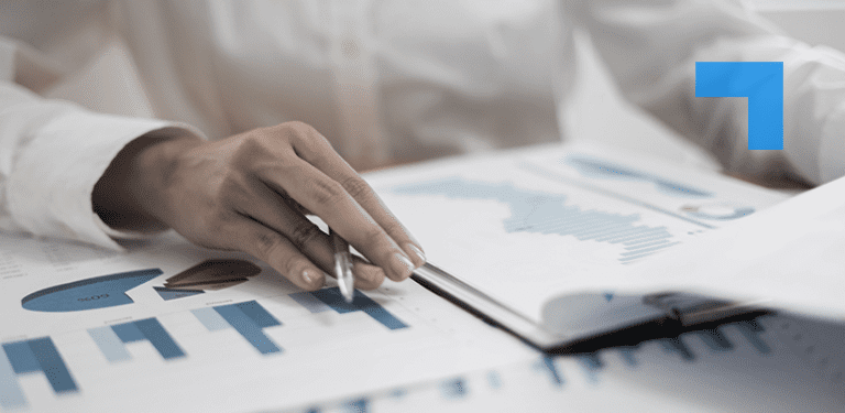 A close-up of a person’s hand holding a pen and pointing at printed financial charts and graphs on a desk, with a notebook partially open nearby.
