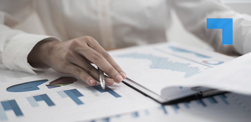 A close-up of a person’s hand holding a pen and pointing at printed financial charts and graphs on a desk, with a notebook partially open nearby.