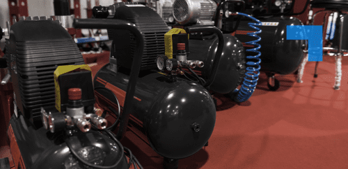 Industrial air compressors displayed on a showroom floor, with pressure gauges and coiled blue air hose visible in a warehouse setting.