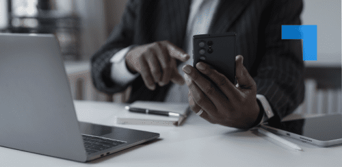 A person in a dark pinstripe suit sits at a desk using a smartphone while working on a laptop. A tablet, stylus and notebook are on the desk.