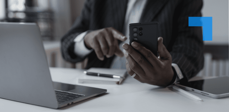 A person in a dark pinstripe suit sits at a desk using a smartphone while working on a laptop. A tablet, stylus and notebook are on the desk.
