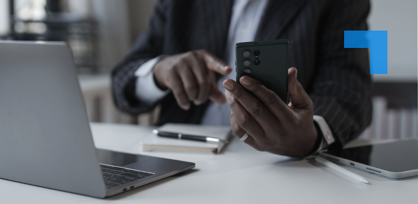 A person in a dark pinstripe suit sits at a desk using a smartphone while working on a laptop. A tablet, stylus and notebook are on the desk.