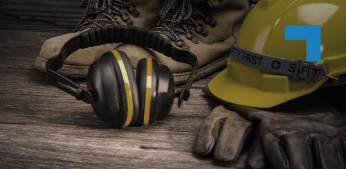 Personal protective equipment arranged on a wooden surface, including a yellow safety hard hat, ear defenders, protective gloves and work boots, representing workplace health and safety in an industrial environment.