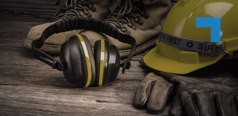 Personal protective equipment arranged on a wooden surface, including a yellow safety hard hat, ear defenders, protective gloves and work boots, representing workplace health and safety in an industrial environment.