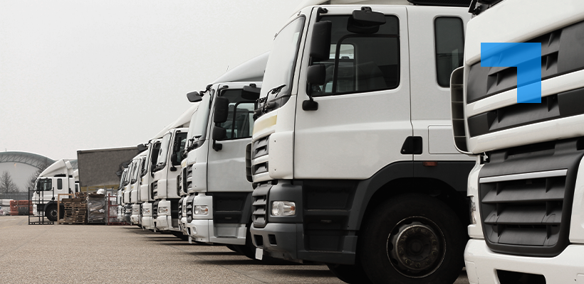 A row of white commercial trucks is parked side by side in an outdoor depot or vehicle yard. The image is taken from a low angle along the line of trucks, emphasizing their size and creating depth as they recede into the distance. Industrial buildings, pallets, and equipment are visible in the background under an overcast sky.