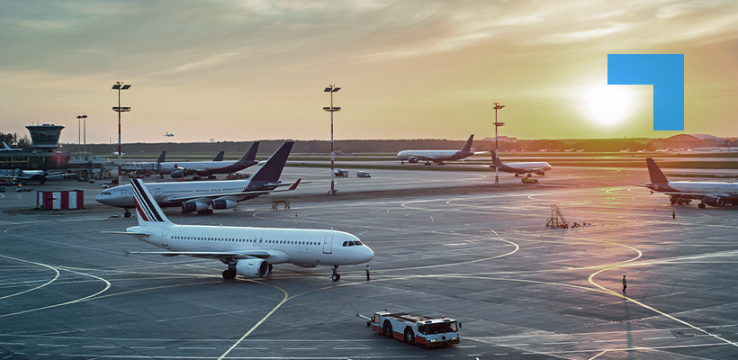 Wide view of an airport apron at sunset, with several commercial aircraft parked or taxiing, ground support vehicles in operation, and terminal buildings visible in the background.