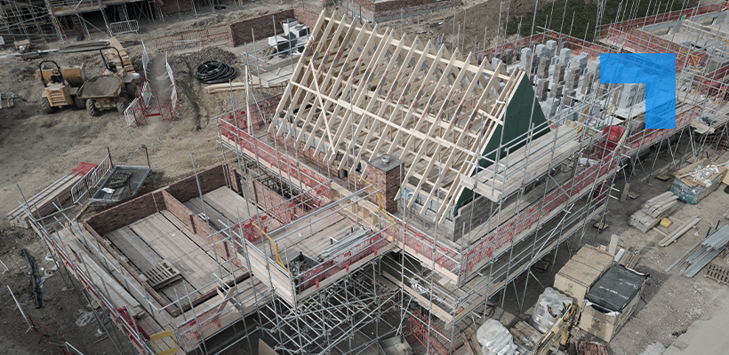 Aerial view of a building under construction, showing roof trusses being installed on a scaffolded structure, with construction materials, machinery and foundation works visible across the site.