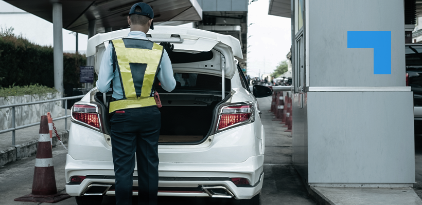 A security officer wearing a high-visibility vest inspects the open trunk of a white car at a checkpoint. Traffic cones line the inspection lane.