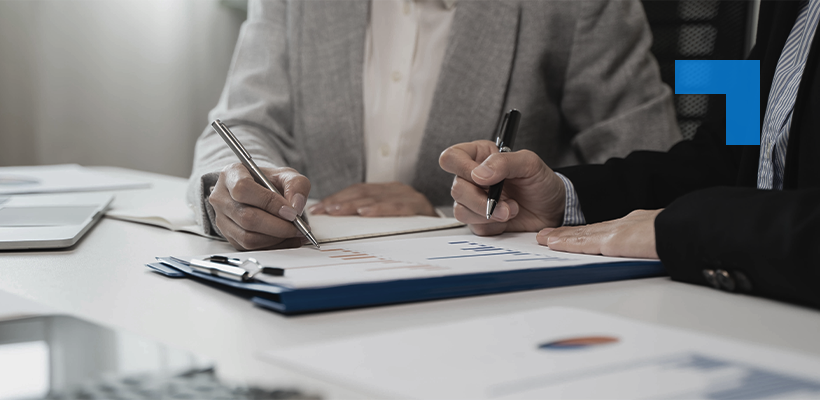 Two professionals seated at a desk, each holding a pen and reviewing printed documents containing charts and data.